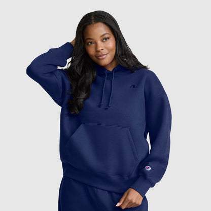 A woman with long dark hair wears a Champion Small C Freshman Powerblend Hoodie in navy blue along with matching sweatpants, standing against a light gray background and smiling as she touches her hair.