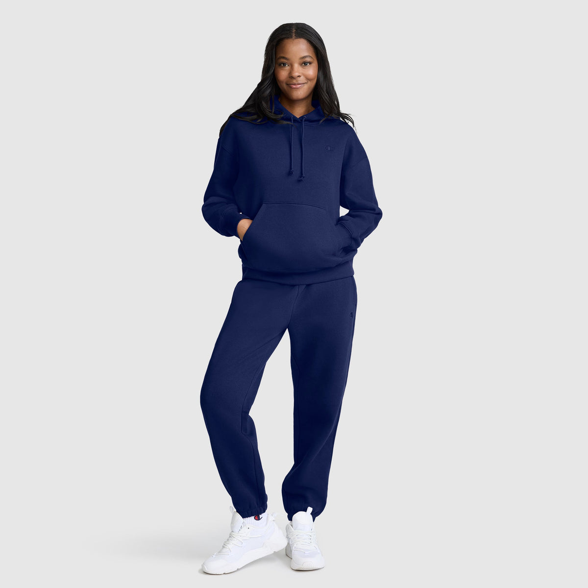 A woman stands smiling against a plain light gray background, wearing the Champion Small C Freshman Powerblend Hoodie paired with matching navy blue sweatpants and white sneakers.