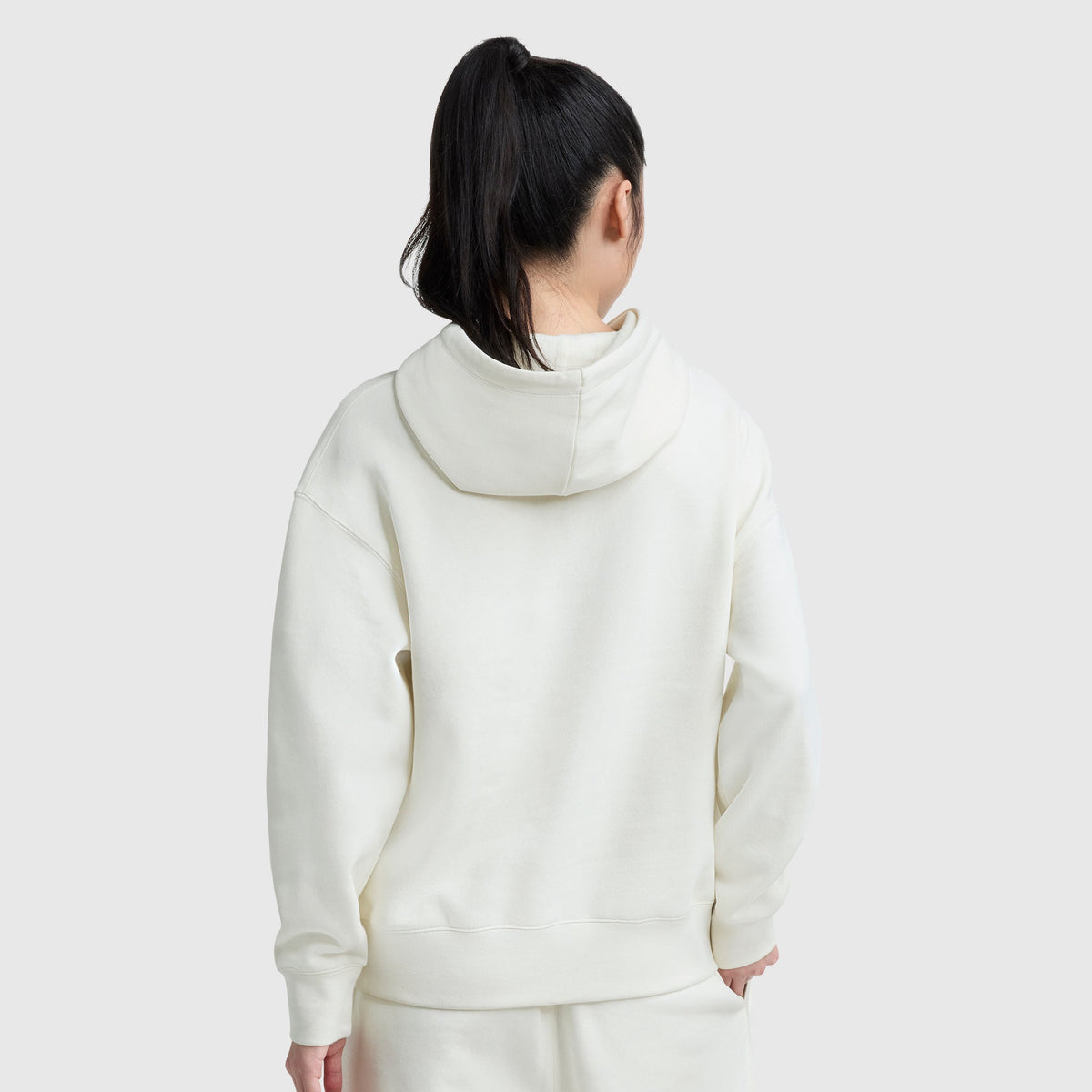 A person with long dark hair in a ponytail is seen from behind, wearing an off-white Champion Small C Freshman Powerblend Hoodie and matching pants against a plain light gray background.
