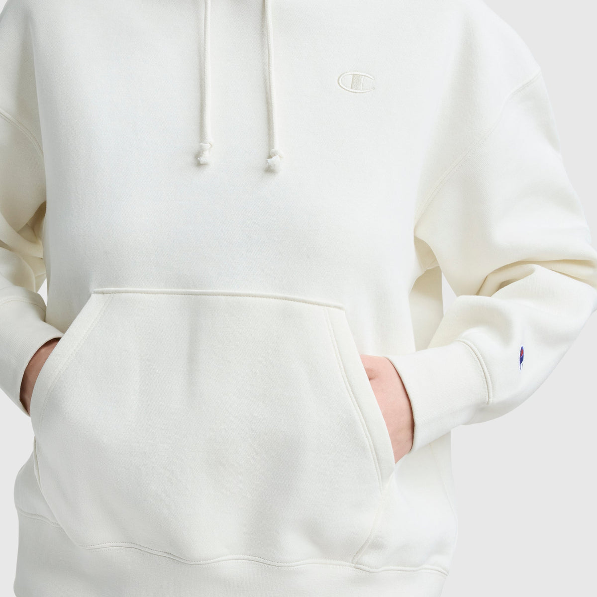 A person wears the Champion Small C Freshman Powerblend Hoodie in off-white, hands tucked in the front pocket. Only their torso and part of the arms are visible. The chest and sleeve display the Champion logo on this midweight fleece hoodie.