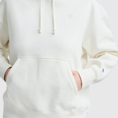A person wears the Champion Small C Freshman Powerblend Hoodie in off-white, hands tucked in the front pocket. Only their torso and part of the arms are visible. The chest and sleeve display the Champion logo on this midweight fleece hoodie.