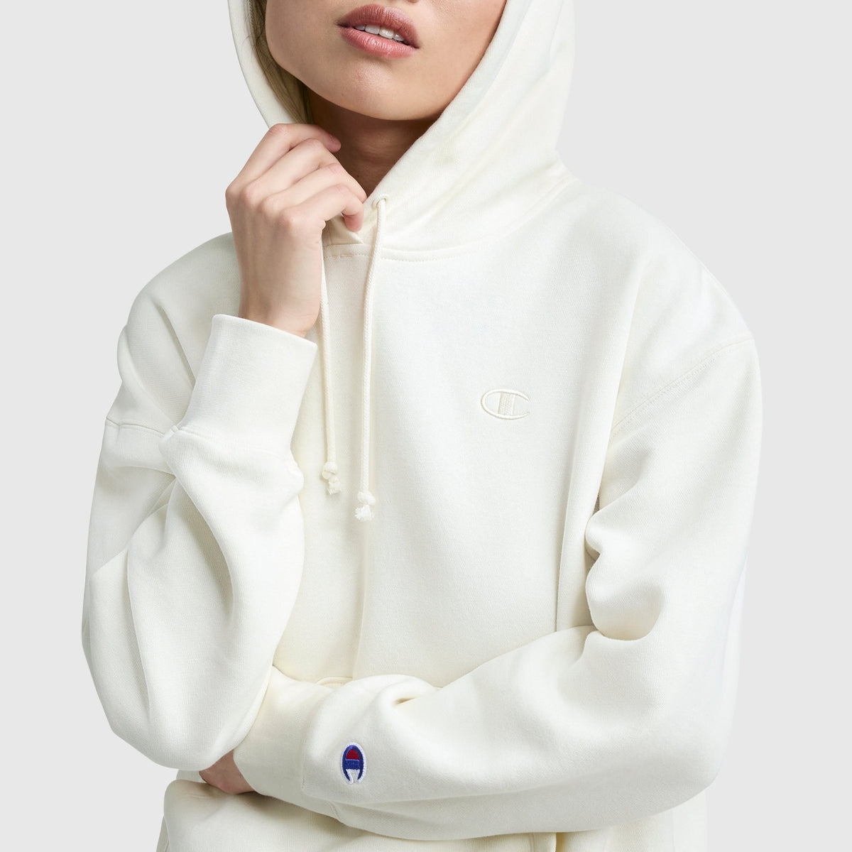 Someone wearing the Champion Small C Freshman Powerblend Hoodie in off-white, hood up and hand on chin. This midweight fleece features a small embroidered logo on the chest and sleeve, set against a plain light gray background.
