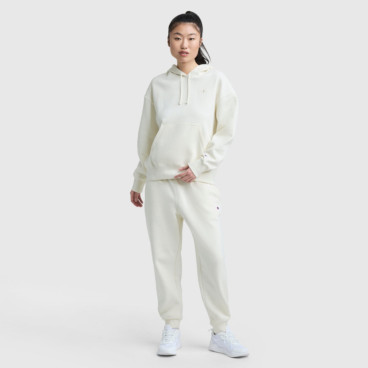 A woman stands against a plain background wearing the Champion Small C Freshman Powerblend Hoodie with matching jogger sweatpants and white sneakers. Her hair is tied back, and she poses casually with one hand in her pocket.
