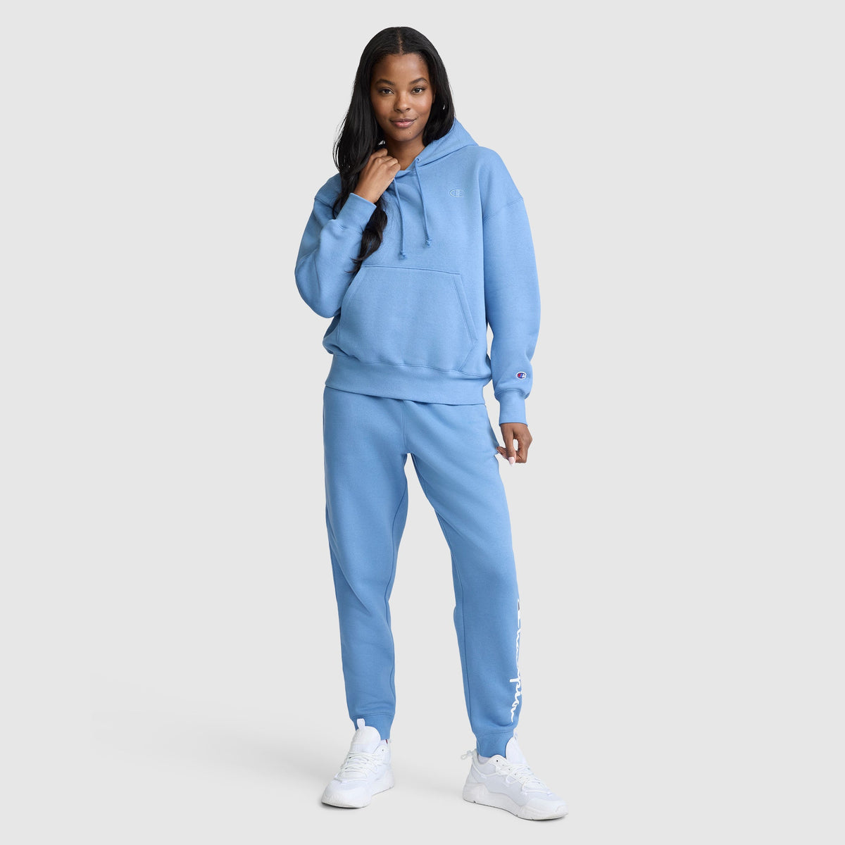 A woman stands confidently wearing a matching light blue Champion Small C Freshman Powerblend Hoodie and sweatpants with white sneakers, posing against a plain white background, effortlessly showcasing Champion style.