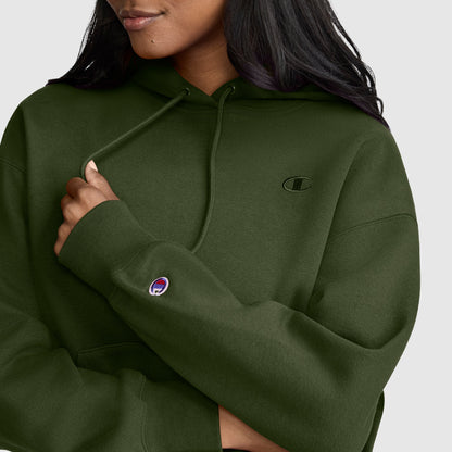 A woman wearing the Champion Small C Freshman Powerblend Hoodie in olive green, featuring a small Champion logo on the chest and a purple logo patch on the sleeve, holds one drawstring against a light gray background.