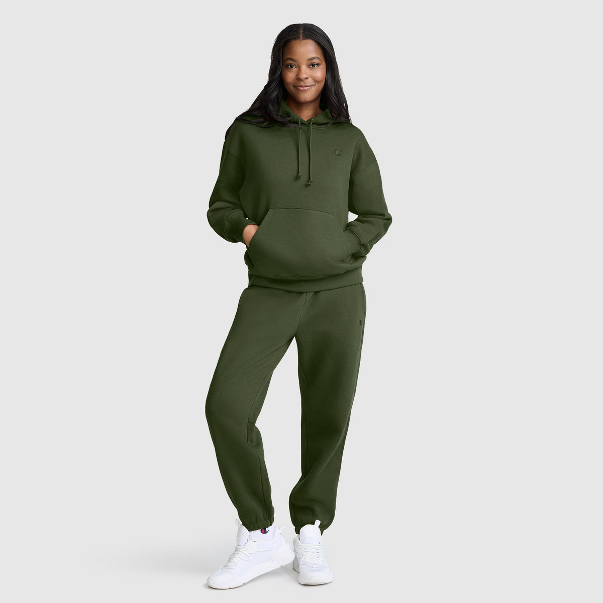 A woman stands smiling with her hands in the pocket of her Champion Small C Freshman Powerblend Hoodie and matching olive green sweatpants, wearing white sneakers against a plain light gray background.