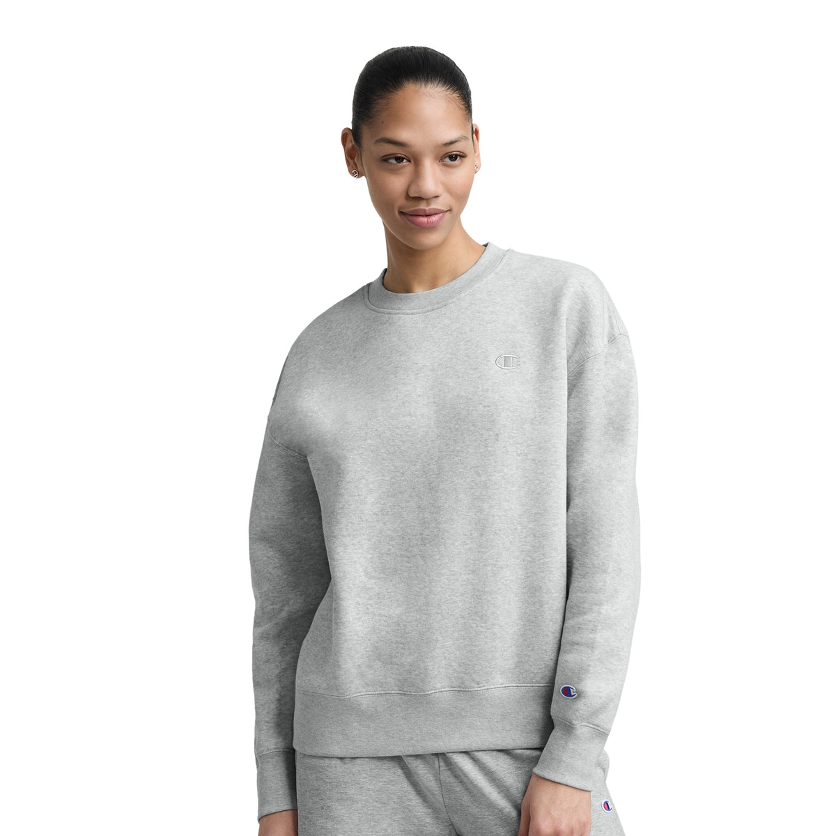 Wearing the Champion Powerblend Crewneck, Small "C," a person in matching light gray sweatpants stands against a white background, looking to the side and smiling in this cozy midweight fleece sweatshirt.