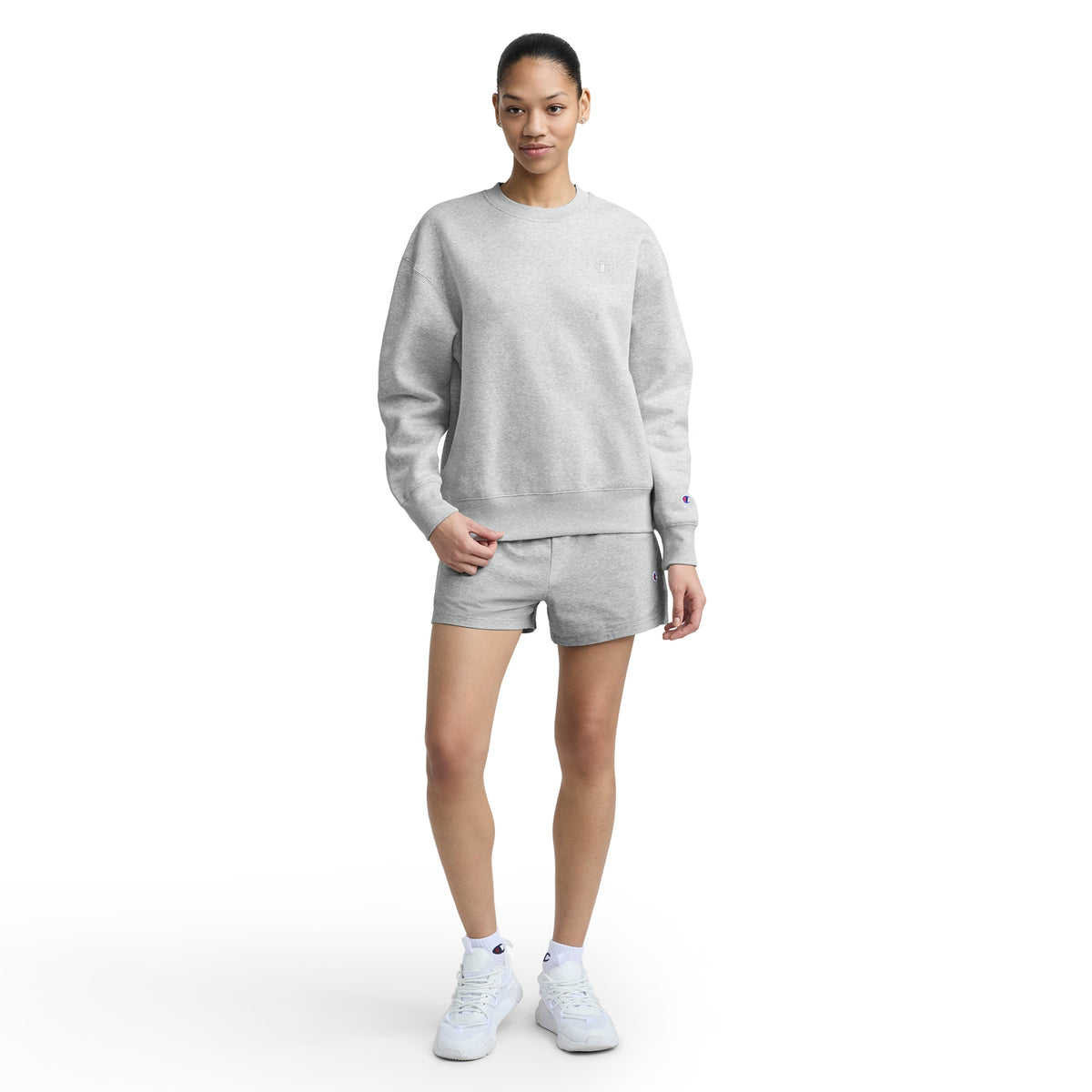 A woman stands against a white background wearing a Champion Powerblend Crewneck, matching shorts, and white sneakers. She has her right hand in her shorts pocket and looks forward with a neutral expression.