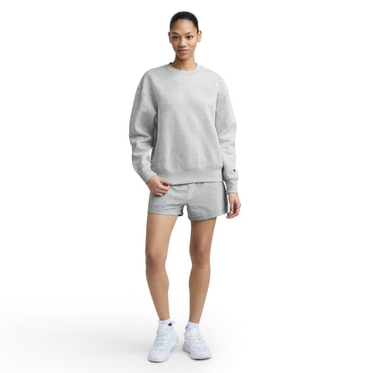 A woman stands against a white background wearing a Champion Powerblend Crewneck, matching shorts, and white sneakers. She has her right hand in her shorts pocket and looks forward with a neutral expression.