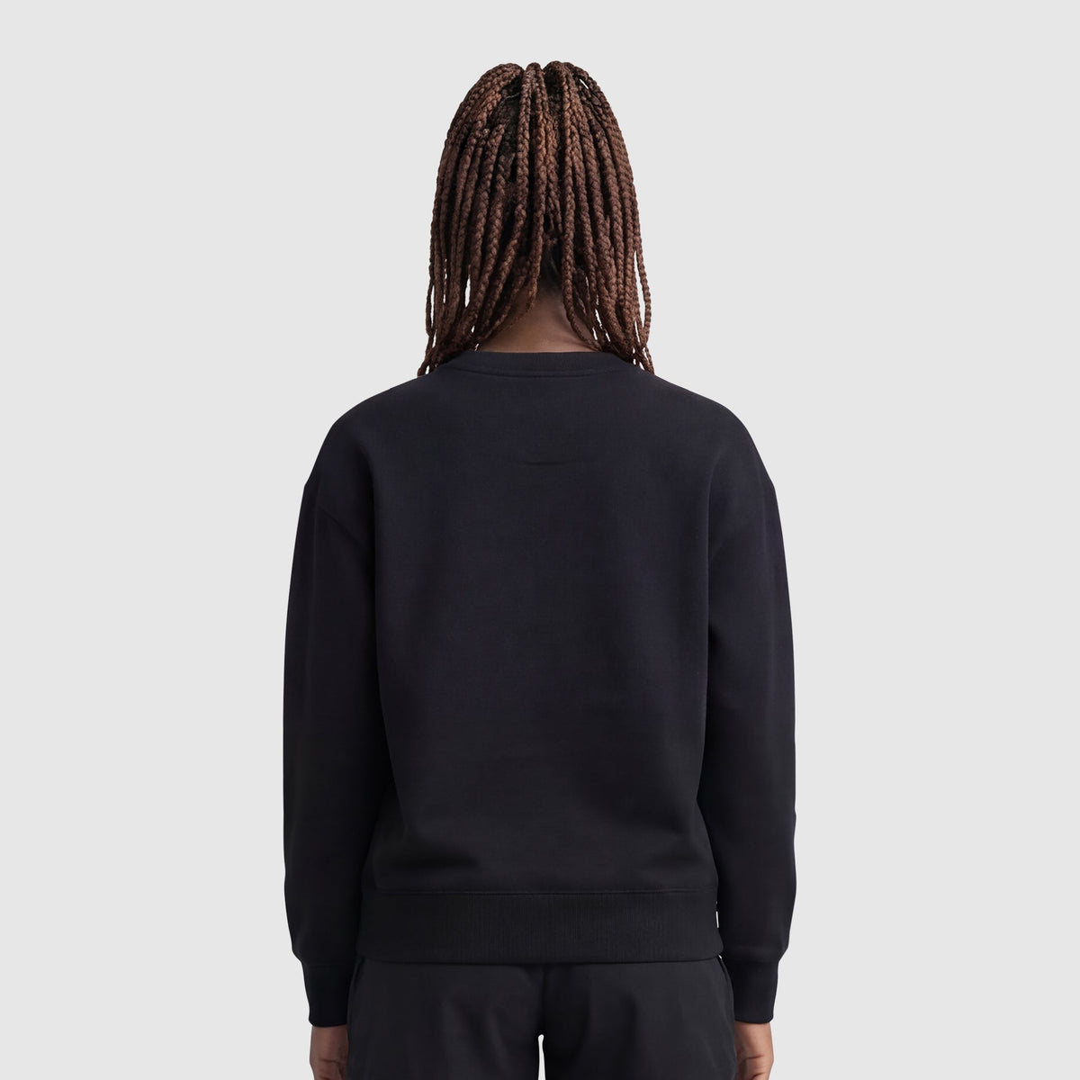 A person with long, braided hair stands with their back to the camera, wearing a Champion Powerblend Crewneck Sweatshirt, Small "C", and black pants against a plain light gray background.
