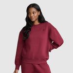 A woman with long dark hair wears a matching maroon Champion Powerblend Crewneck, Small “C”, and sweatpants, standing against a plain light gray background with one hand on her hip and a slight smile.