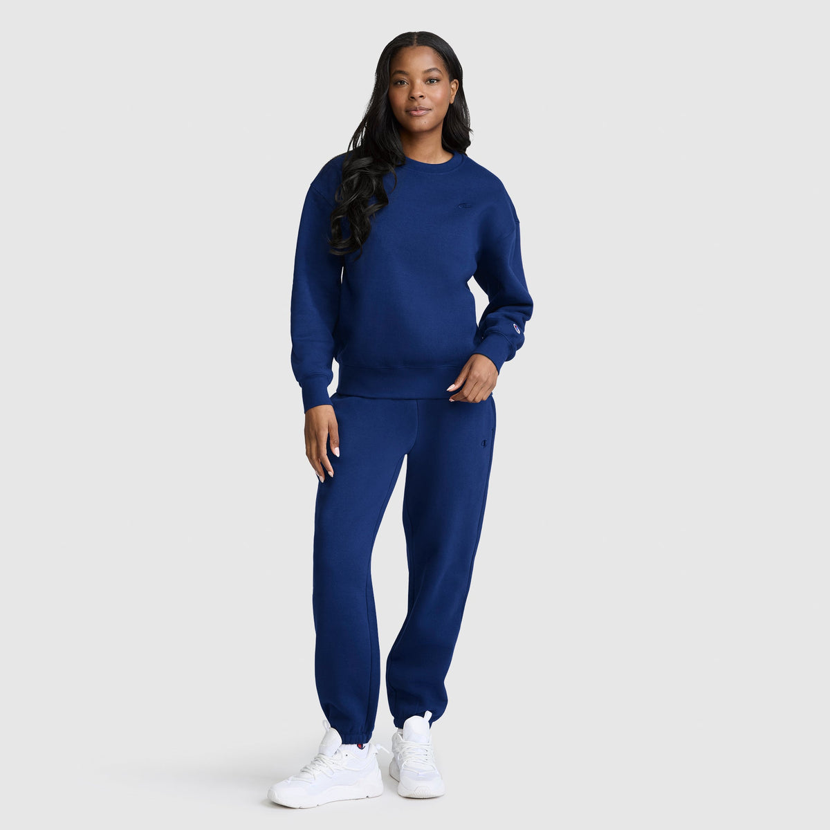 A woman stands wearing the Champion Powerblend Crewneck, Small “C” sweatshirt paired with matching dark blue fleece sweatpants and white sneakers, posing against a plain light gray background.