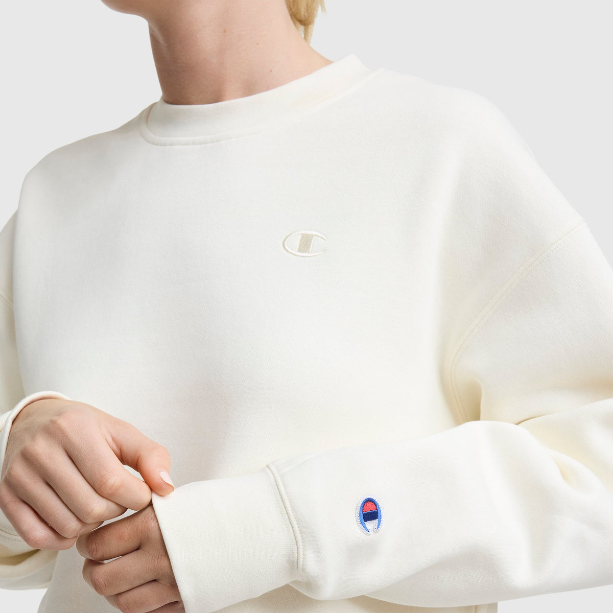 A person wearing a Champion Powerblend Crewneck, featuring an off-white color, midweight fleece, and small "C" logos embroidered on the chest and sleeve, appears with their head partially out of frame.