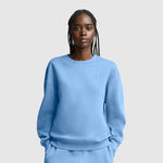 A woman with braided hair wears a light blue Champion Powerblend Crewneck, Small “C”, and matching pants as she stands against a plain light gray background with her hands in her pockets.