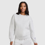 A woman stands against a plain light background, looking at the camera with a slight smile, dressed in a light gray Champion Powerblend Crewneck Sweatshirt with an embroidered script logo and matching sweatpants. This cozy fleece set is an ideal choice for anyone who values comfort and timeless style from Champion's women's collection.