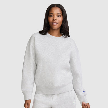 A woman stands against a plain light background, looking at the camera with a slight smile, dressed in a light gray Champion Powerblend Crewneck Sweatshirt with an embroidered script logo and matching sweatpants. This cozy fleece set is an ideal choice for anyone who values comfort and timeless style from Champion's women's collection.