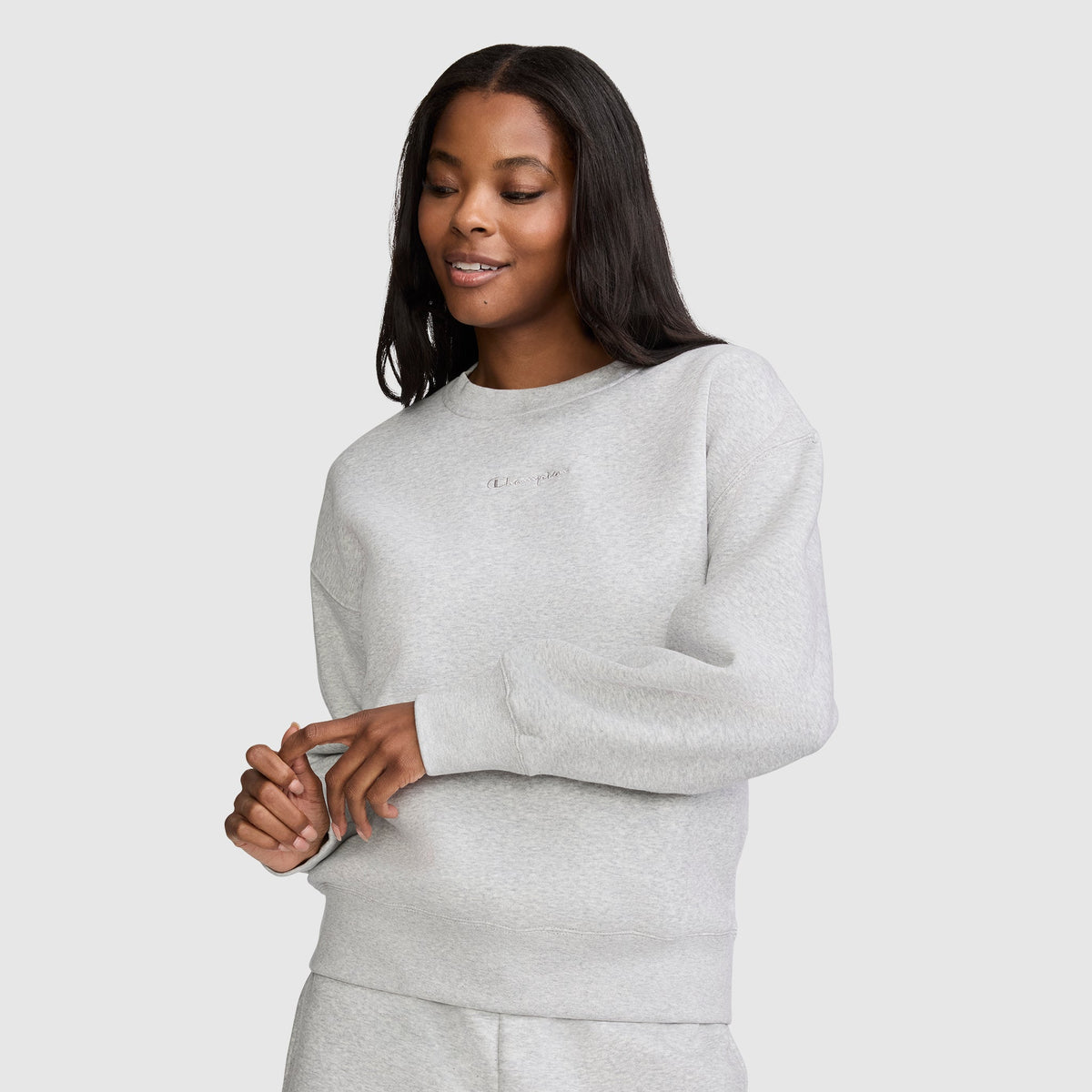 A woman with long dark hair wears the Champion Powerblend Crewneck Sweatshirt with Embroidered Script Logo and matching pants, standing against a plain light background and smiling softly with her hands gently clasped in front of her. Crafted for comfort and style, the Powerblend fleece sets this Champion sweatshirt apart in women's fashion.