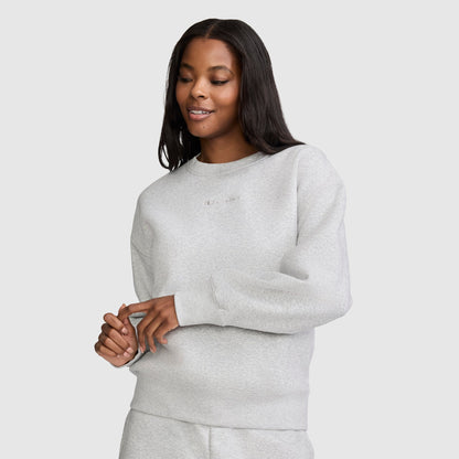 A woman with long dark hair wears the Champion Powerblend Crewneck Sweatshirt with Embroidered Script Logo and matching pants, standing against a plain light background and smiling softly with her hands gently clasped in front of her. Crafted for comfort and style, the Powerblend fleece sets this Champion sweatshirt apart in women's fashion.