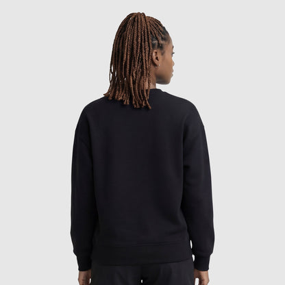 A person with braided hair, seen from behind, is wearing a black Champion Powerblend Crewneck Sweatshirt with an embroidered script logo and black pants, standing against a light gray background.