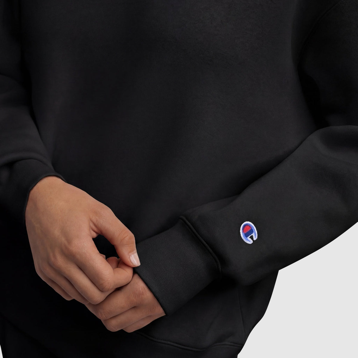 A woman adjusts the sleeve of a black Champion Powerblend Crewneck Sweatshirt, Embroidered Script Logo, showing a small circular red, white, and blue logo near the wrist against a plain light background.