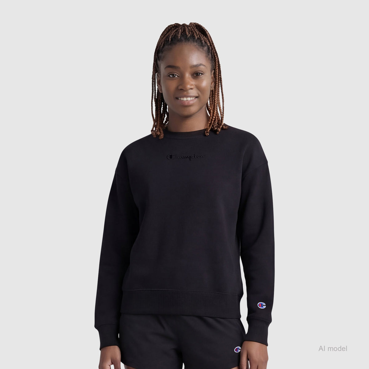 A woman with braided hair wears the Champion Powerblend Crewneck Sweatshirt with embroidered script logo and black shorts, standing against a plain light gray background, facing forward and smiling softly.