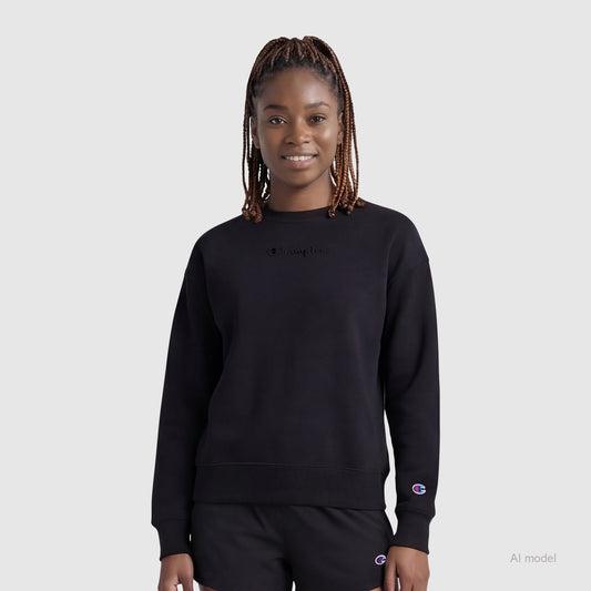 A woman with braided hair wears the Champion Powerblend Crewneck Sweatshirt with embroidered script logo and black shorts, standing against a plain light gray background, facing forward and smiling softly.