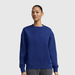 A person stands against a plain white background, smiling slightly with hands relaxed at their sides, wearing a Champion Powerblend Crewneck Sweatshirt with an embroidered script logo in blue and gray pants.
