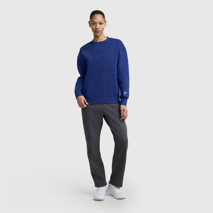 A person stands against a light gray background wearing a Champion Powerblend Crewneck Sweatshirt with an embroidered script logo in blue, paired with gray pants and white sneakers, one hand in their pocket and a slight smile on their face.