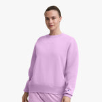 A woman wearing a light purple Champion Powerblend Crewneck Sweatshirt with an embroidered script logo and matching pants stands against a plain white background, looking forward with a slight smile.