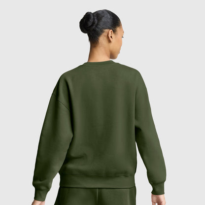 A woman with her hair in a bun, dressed in an olive green Champion Powerblend Crewneck Sweatshirt with an embroidered script logo and matching pants, stands with her back to the camera against a plain light gray background.
