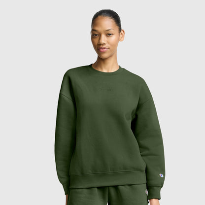 A woman dressed in an oversized dark green sweatshirt and matching pants stands against a plain light gray background, gazing at the camera with a neutral expression. Her relaxed outfit captures the essence of women’s fashion sweatshirts, similar to the Champion Powerblend Crewneck Sweatshirt with Embroidered Script Logo, renowned for its soft fleece comfort.