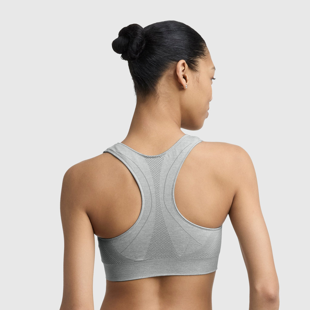 A woman with dark hair in a bun is shown from the back wearing the Champion Infinity Racerback Sports Bra, a light gray seamless sports bra, against a plain light background.