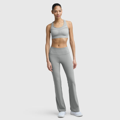 A woman stands against a plain background, smiling softly in a grey Champion Infinity Racerback Sports Bra with moderate support, paired with matching high-waisted flared leggings and white sneakers. One hand rests by her side.