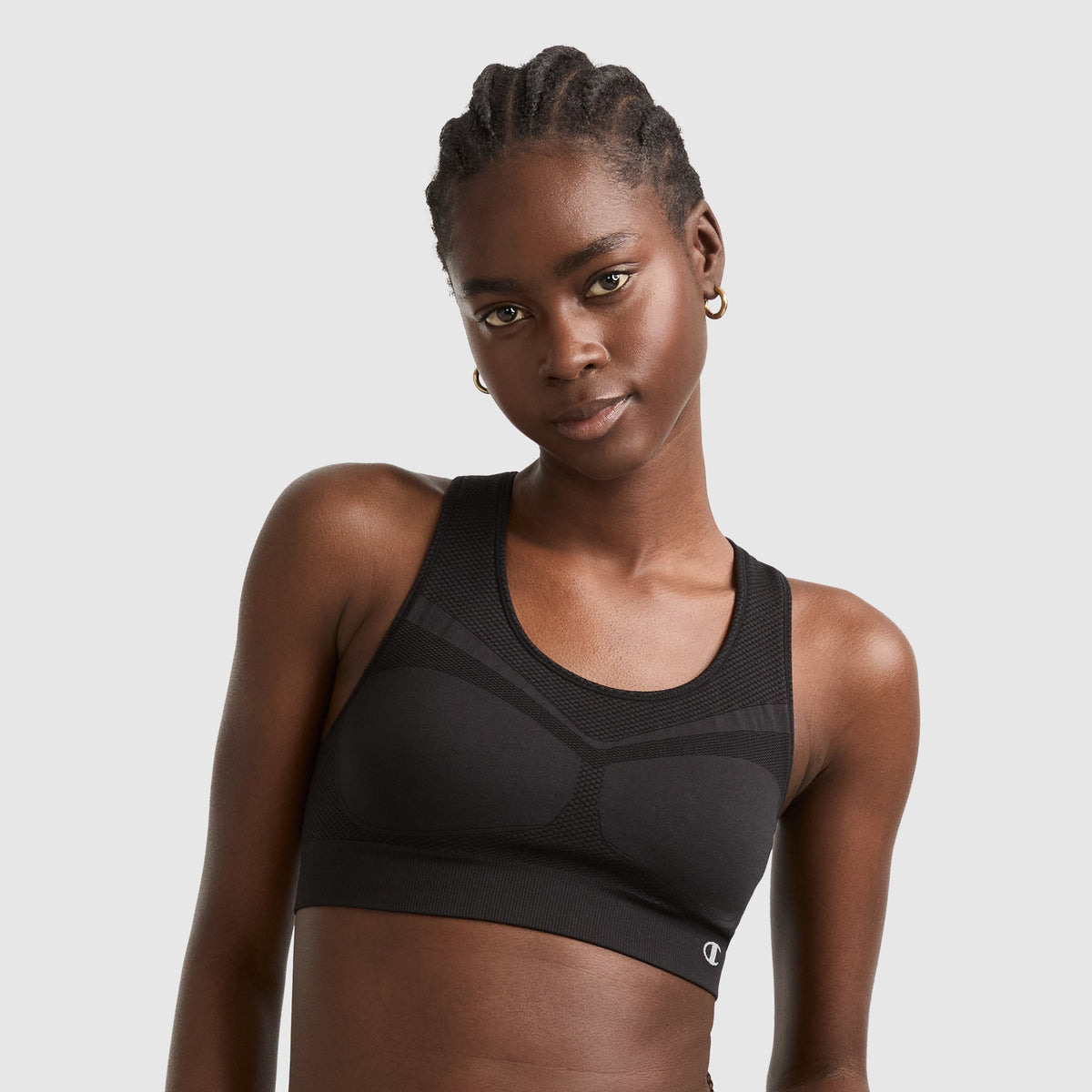 A woman with braided hair wears a black sports bra and gold hoop earrings, standing against a plain light gray background, looking into the camera with a slight smile.