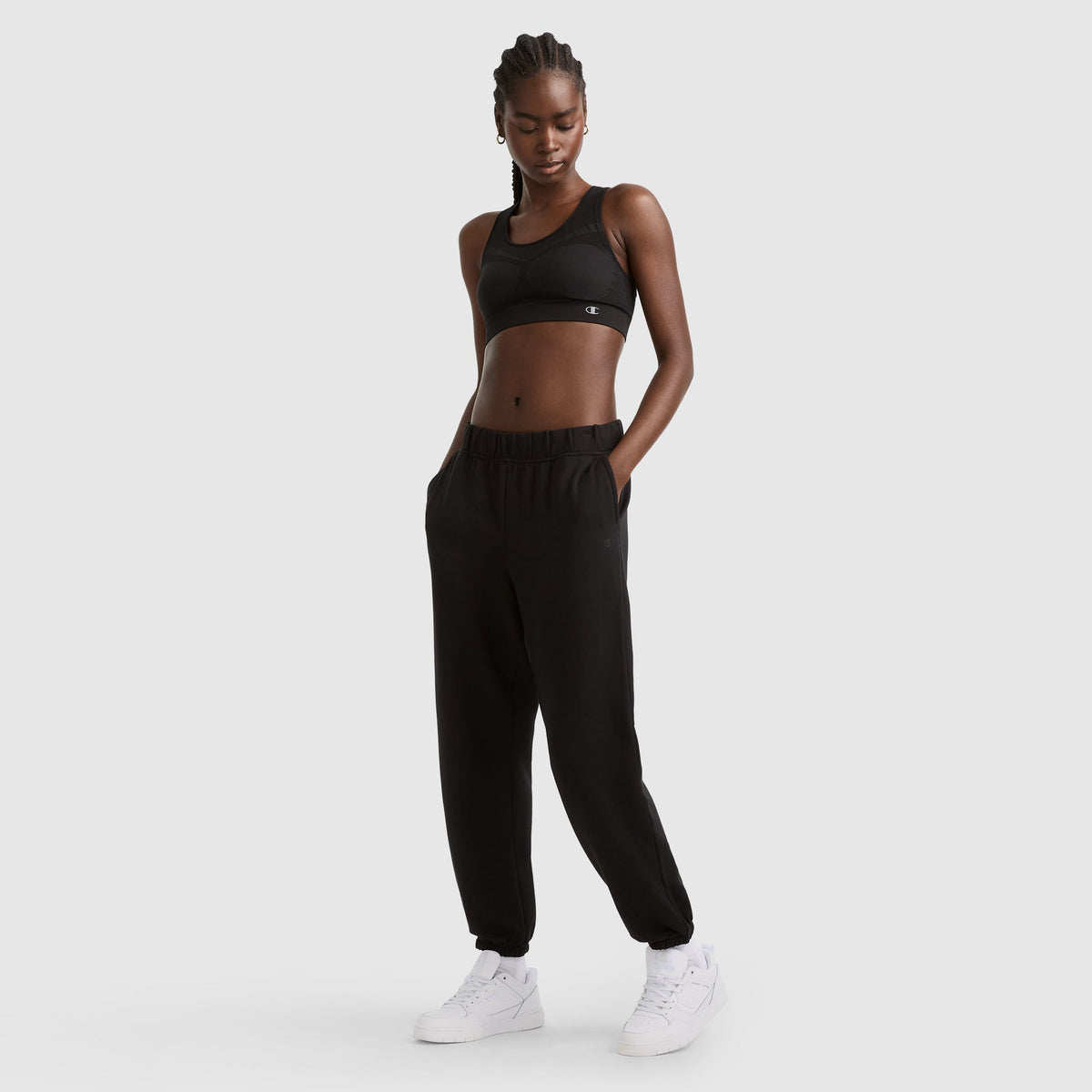 A woman with braided hair wears a black sports bra, black jogger pants, and white sneakers. She stands with hands in her pockets against a plain light gray background.