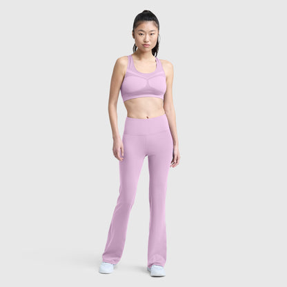 A person stands facing forward, wearing the Champion Seamless Racerback Sports Bra in light purple with moderate support and matching flared leggings, paired with white sneakers against a plain light gray background.