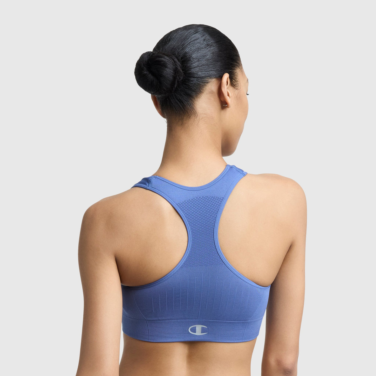A woman with dark hair in a neat bun is seen from the back wearing the Champion Infinity Racerback Sports Bra in blue, featuring a small logo near the hem against a plain light gray background.