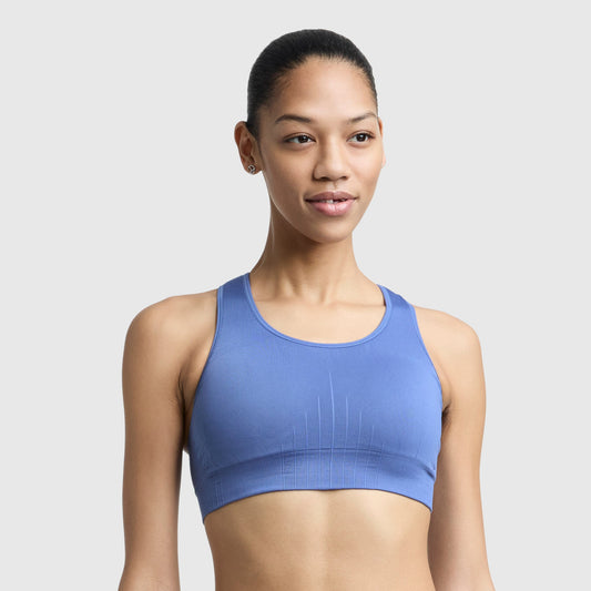 A woman with dark hair pulled back wears a blue Champion Infinity Racerback Sports Bra and small stud earrings, standing against a plain light gray background and gazing slightly to the side.
