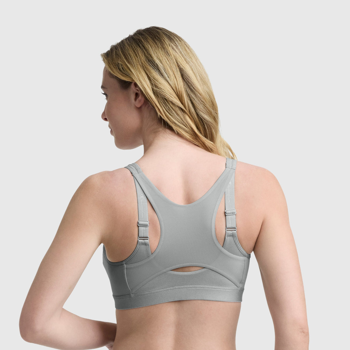 A woman with long blonde hair is shown from the back, wearing the Champion Motion Control Zip Sports Bra in grey, featuring adjustable straps and a small keyhole cutout below the shoulder blades. The background is plain light grey.