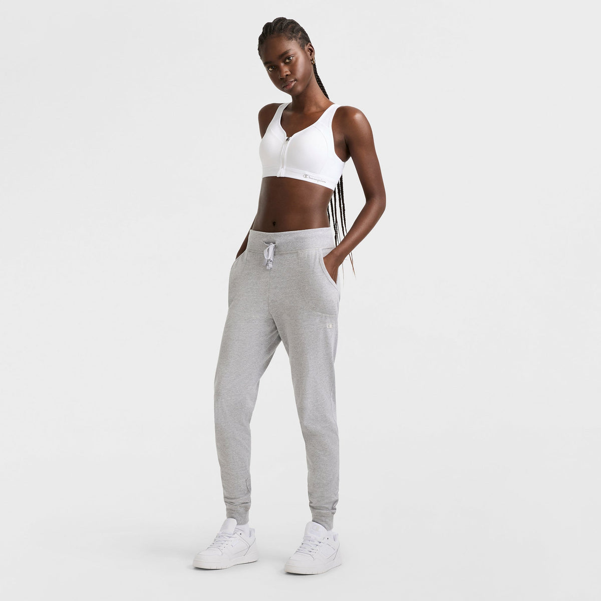 A woman stands confidently with her hands in the pockets of light gray joggers, wearing a Champion Motion Control Zip Sports Bra, Wirefree, Script Logo in white and white sneakers, against a plain white background.