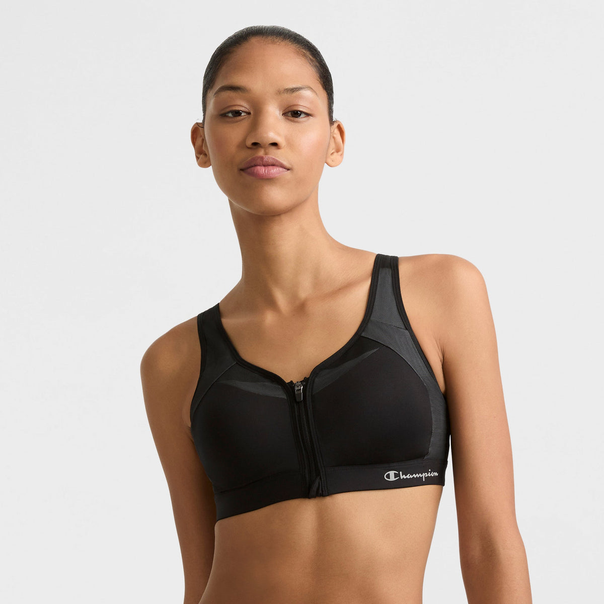 A woman models the Champion Motion Control Zip Sports Bra, Wirefree, with a script logo on the band, standing against a plain light background and looking confidently at the camera.