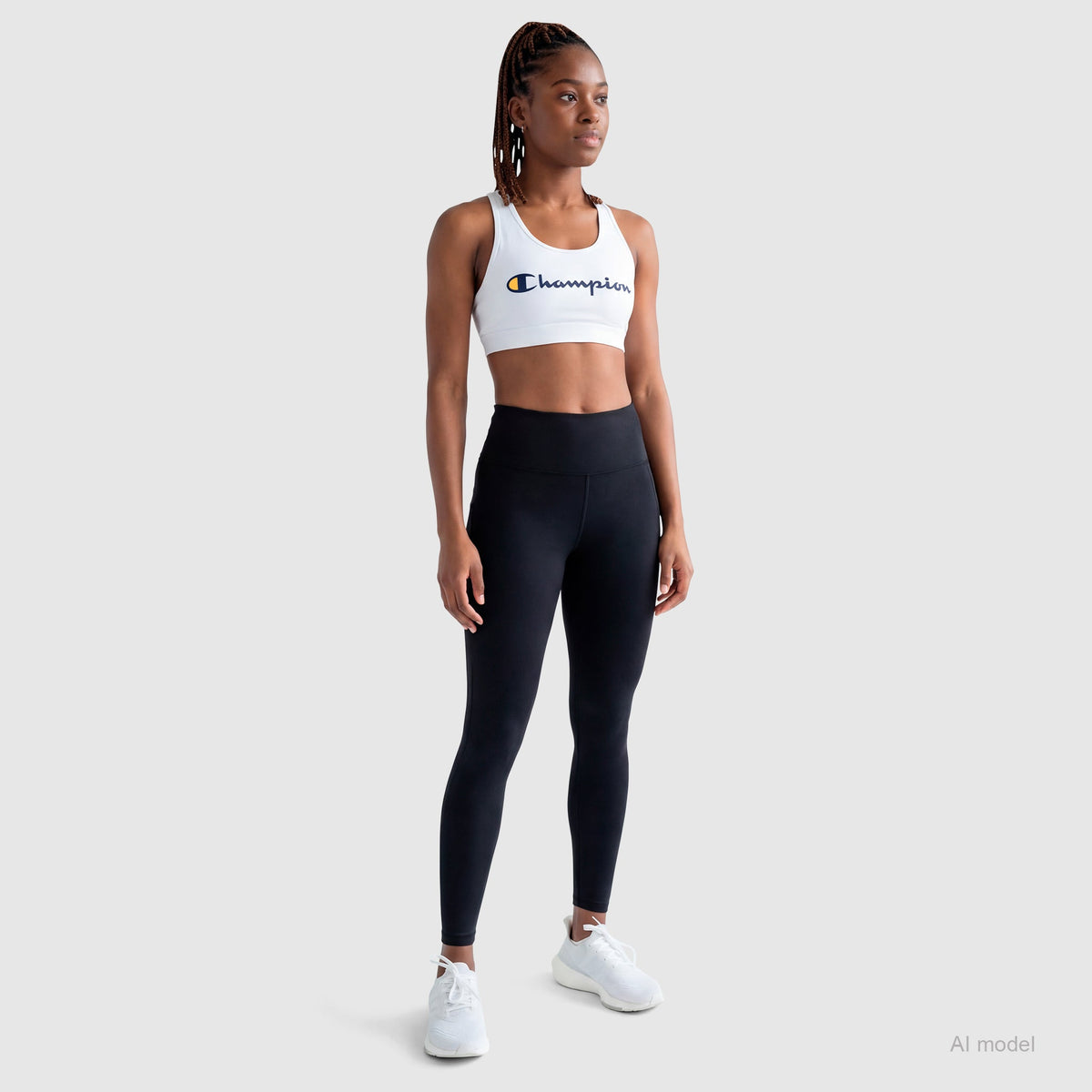 A woman stands against a light gray background wearing the Champion Sports Bra with Script Logo, black high-waisted leggings, and white sneakers. She has braided hair and a confident, relaxed posture. The bra is made from soft cotton-spandex jersey.
