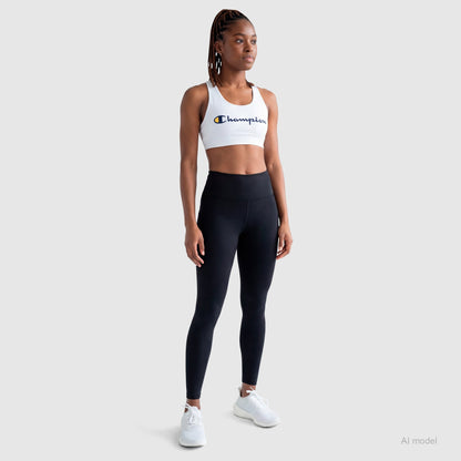 A woman stands against a light gray background wearing the Champion Sports Bra with Script Logo, black high-waisted leggings, and white sneakers. She has braided hair and a confident, relaxed posture. The bra is made from soft cotton-spandex jersey.