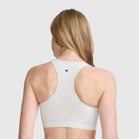A woman with long blonde hair is shown from behind against a plain light background, wearing the Champion Sports Bra, Script Logo—a white cotton-spandex athletic bra with a small Champion logo on the back.