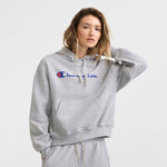 A person wears the Champion Powerblend Crewneck Sweatshirt, Script Logo, in light gray with blue and red branding, standing against a white background and looking at the camera while touching their hair.