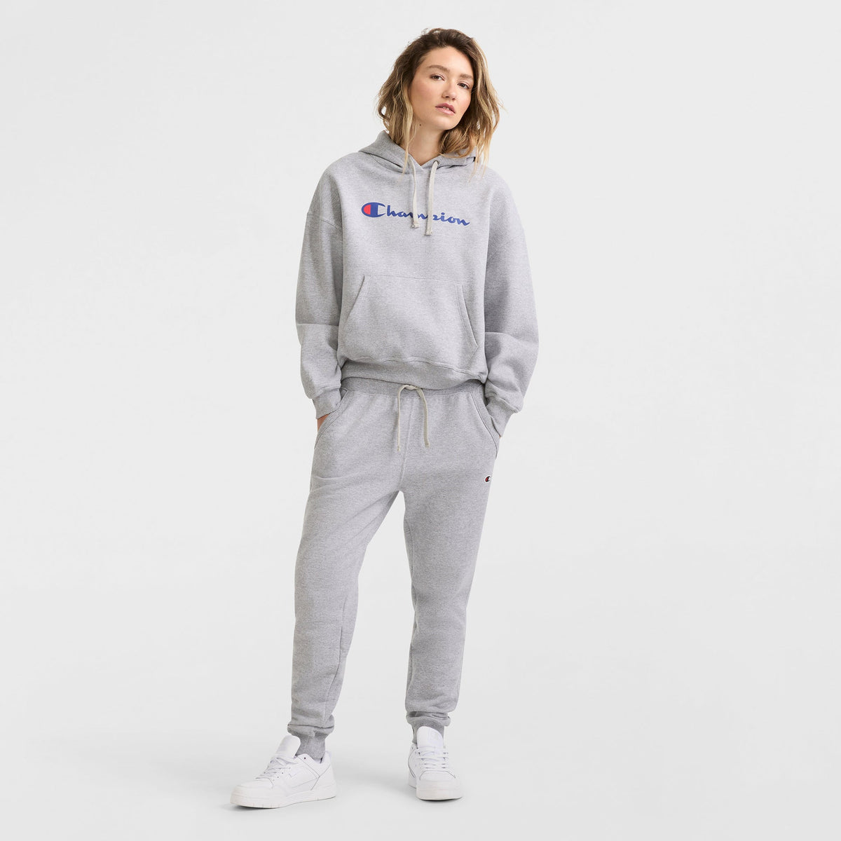 A person stands in a Champion Powerblend Crewneck Sweatshirt with Script Logo, styled in light gray with matching sweatpants and white sneakers. Their shoulder-length hair and relaxed pose are set against a plain white background.