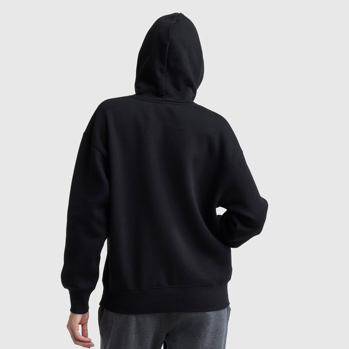 A person wearing a Champion Powerblend Relaxed Hoodie in black and gray pants stands with their back to the camera, hood up and one hand on their hip against a plain, light-colored background.