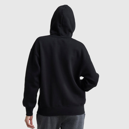 A person wearing a Champion Powerblend Relaxed Hoodie in black and gray pants stands with their back to the camera, hood up and one hand on their hip against a plain, light-colored background.