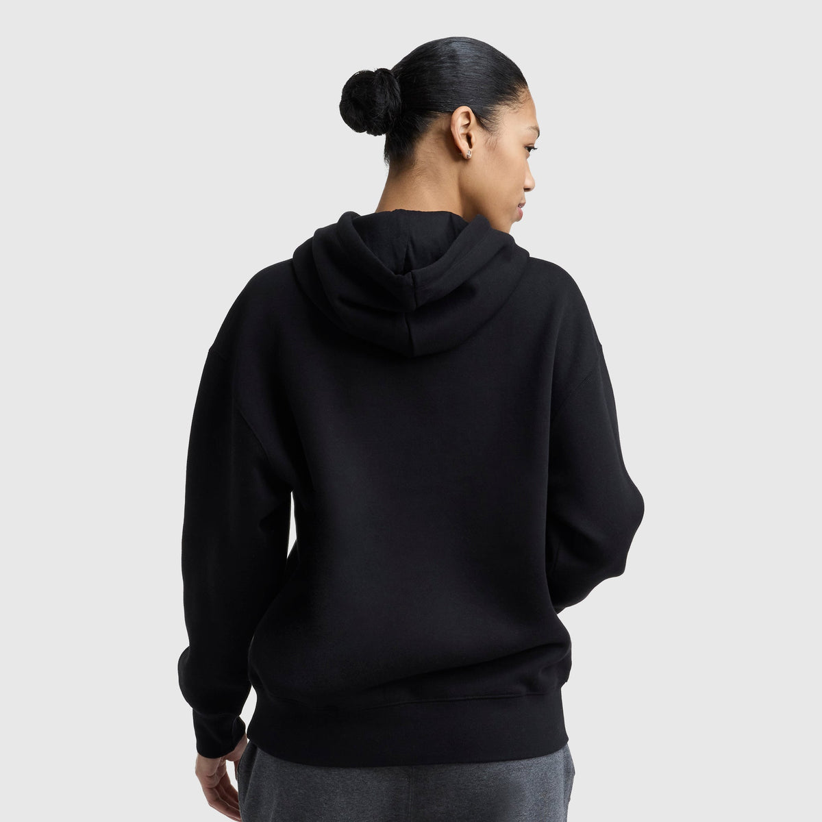 A person with dark hair in a bun is shown from the back wearing a black Champion Powerblend Relaxed Hoodie and gray pants against a plain light background.