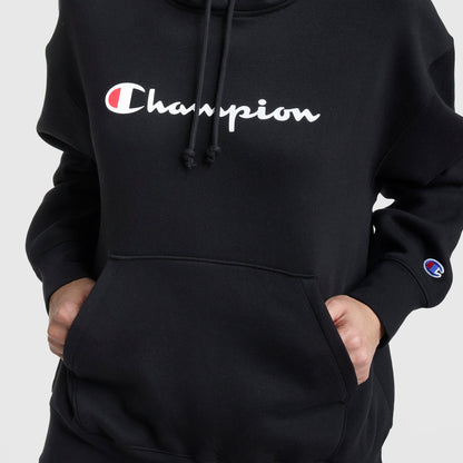A person wears a black Champion Powerblend Relaxed Hoodie with hands in the front pocket. The chest and sleeve feature the white Champion logo with a red and blue "C." The background is plain and light-colored.